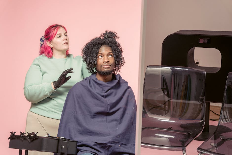 Skilled female hairdresser touching Afro hair of African American male client while sitting near professional supply on pink background in studio