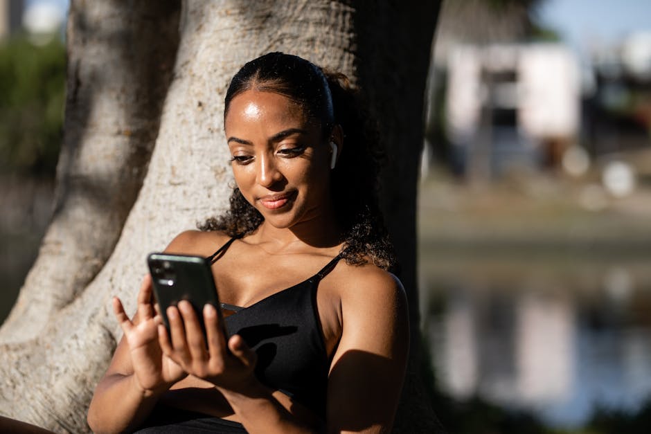 Smiling woman in activewear leans against tree while using smartphone outdoors.
